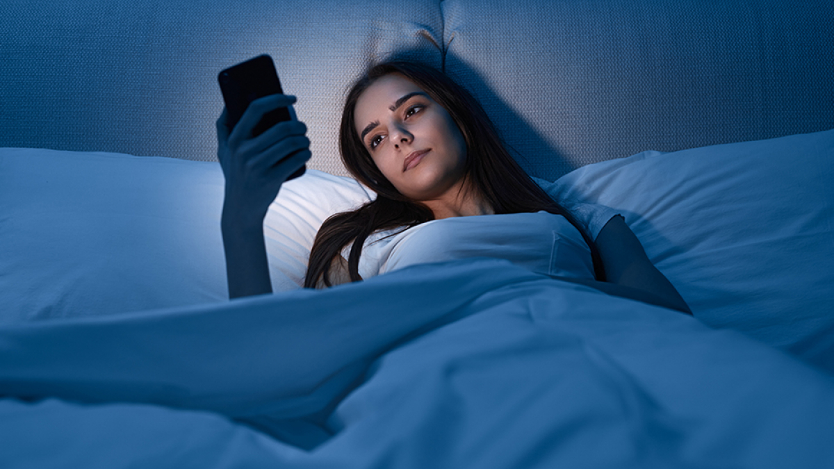 Young woman watching video on cellphone while lying on bed in dark bedroom at night